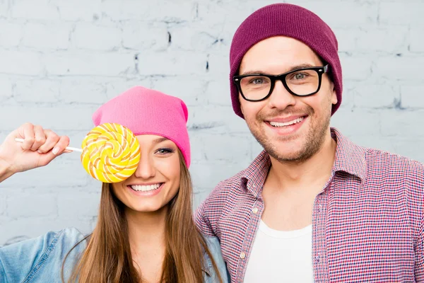 Portrait of happy young comic couple in hats holding lollipop