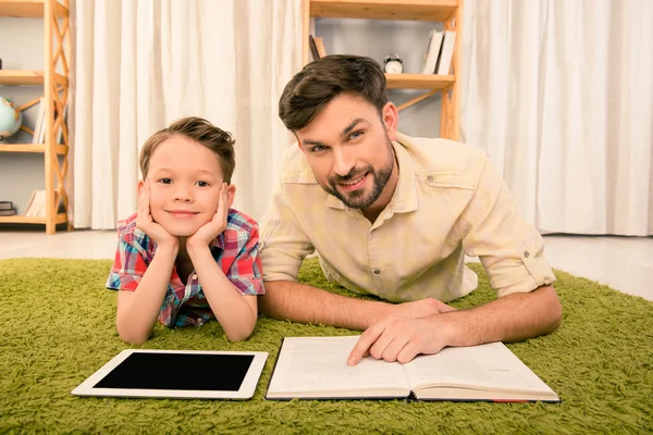 Father and son having fun and lying on carpet with book and tabl