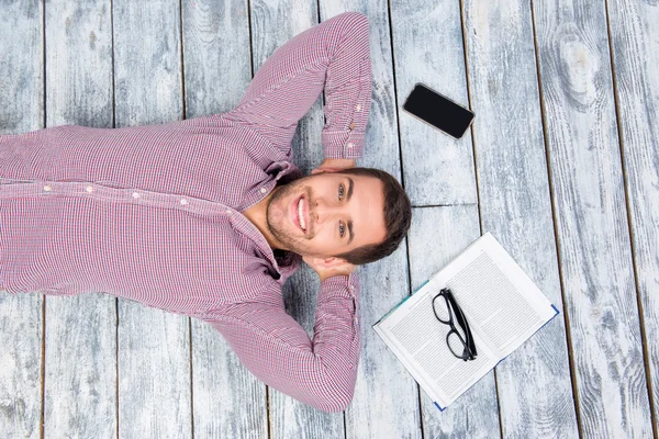 Top view photo of smiling man  lying on the floor with book, phone and glasses