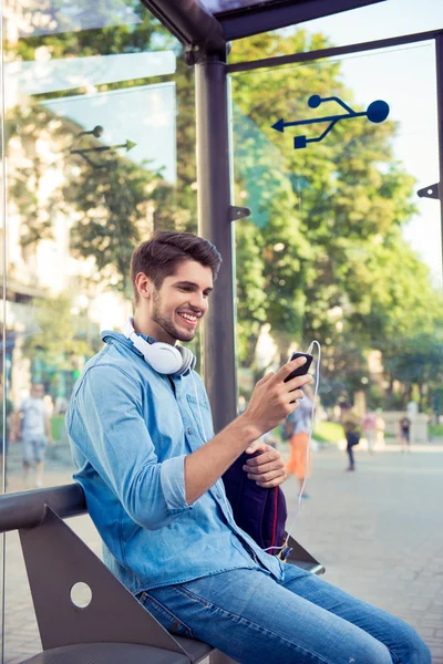 Cheerful young man sitting at bus stop and   waiting