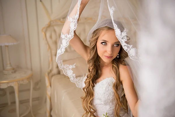 Beautiful bride in her bright bedroom sitting on a luxurious white bed and holding her beautiful long veil veil