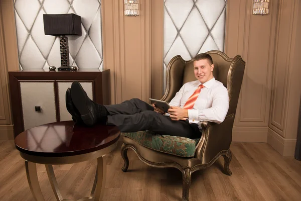 Confidence handsome businessman in chic interior. Successful young man in a business suit sitting on the armchair with his legs crossed and smiling