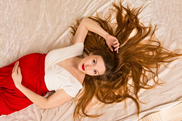 Incredibly beautiful pregnant woman lying on a bed in a red dress with white bodice. her lips painted red lipstick. girl lying on white bed, spreading her long red hair