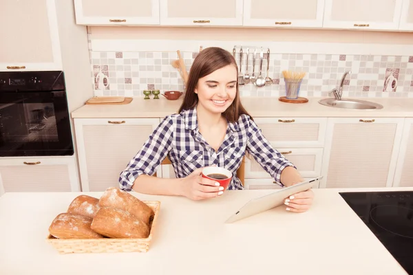 Smiling girl with cup of tea reading news on tablet in kitchen