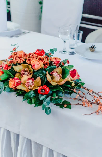 Wedding Banquet in autumn style. The composition of red, orange, yellow, and green, standing on a white table in the area of wedding party. Orchid, rose, eucalyptus, greenery flowers, branches and dec