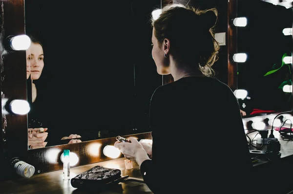 Young woman applying make up, looking herself reflection in mirror with bulbs at dressing in dark interior room. Girl applying cosmetic with brushes, tonal cream.