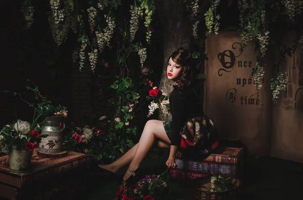 Magic portrait of romantic beautiful girl with wavy hair, red lips, art dress, holding rose flower, sitting on books. Female in scenery of Alice in Wonderland. Fashion fairy tale about princess, walki