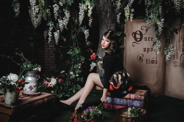 Magic portrait of romantic beautiful girl with wavy hair, red lips, art dress, holding rose flower, sitting on books. Female in scenery  Alice in Wonderland. Fashion fairy tale about princess, walking
