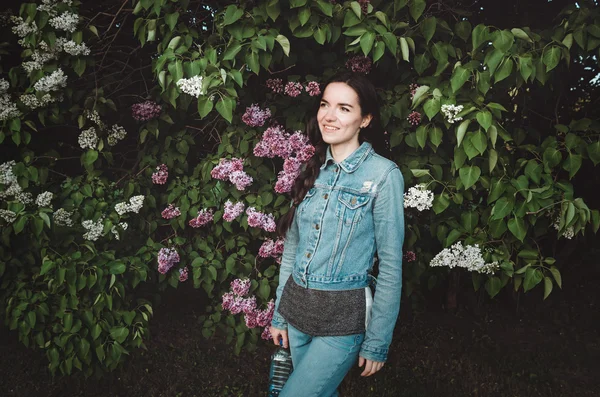 Portrait of a beautiful smiling, young woman outdoor with blossom purple lilac flowers in  spring garden. Attractive female in casual jeans clothes, holding  bottle  water.  concept, nature and care.