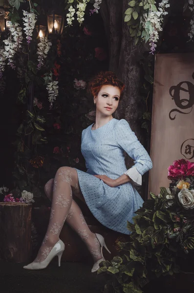 Portrait of romantic redhead young woman with wavy hairstyle, blue vintage dress, white elegant shoes, lace tights, sitting on retro suitcase surrounded blooming floral magic garden  street light