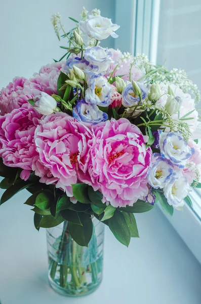 Beautiful bouquet of pink peonies, roses, eustoma flowers in vase on white window sill, cream color background with sun light. Spring or summer lovely . Fresh floral, home decor. Pastel colors purple