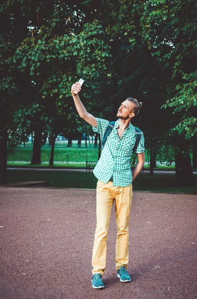 I love selfie. Outdoor summer lifestyle portrait of young bearded man holding camera and making . Smiling hipster guy in shirt walking in city park taking a photo with Smartphone. Modern life, social
