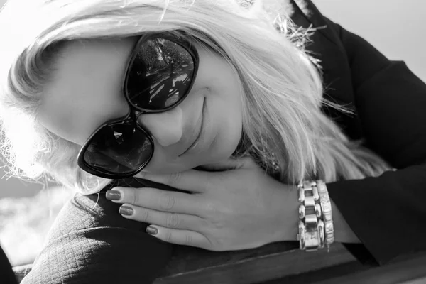 Portrait of a young beautiful woman with blonde hair. She is wearing black glasses and black jacket. Light smile. Discrete make-up. Black and white photo.
