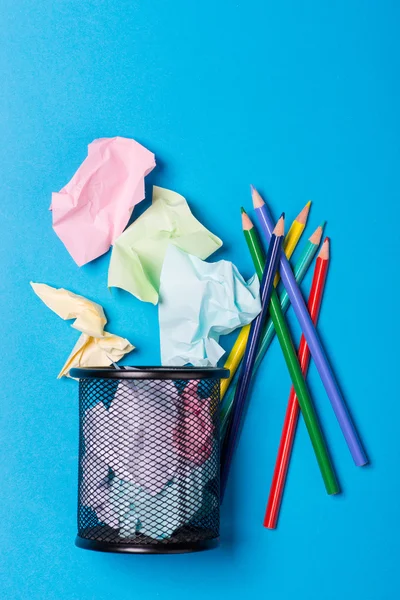 Office trash bin with pencils and crumpled color paper over the blue background
