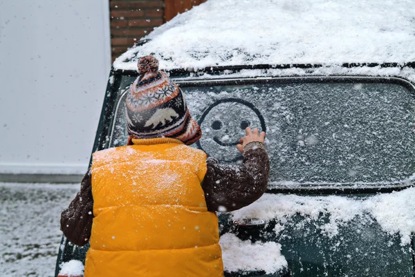 Boy drawing smiley face in snow, on car window.