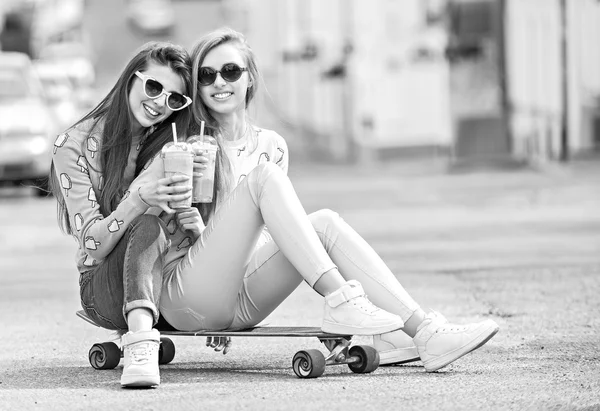 Beautiful young girls hipster girlfriends posing with a skateboard seat on skate, street fashion lifestyle in sunglasses. Keep cocktail and smiling