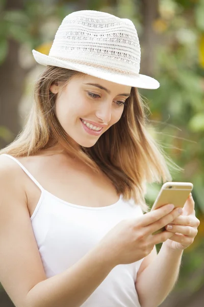 Portrait of a smiling young woman wearing a hat and standing in a garden looking at her cell phone.