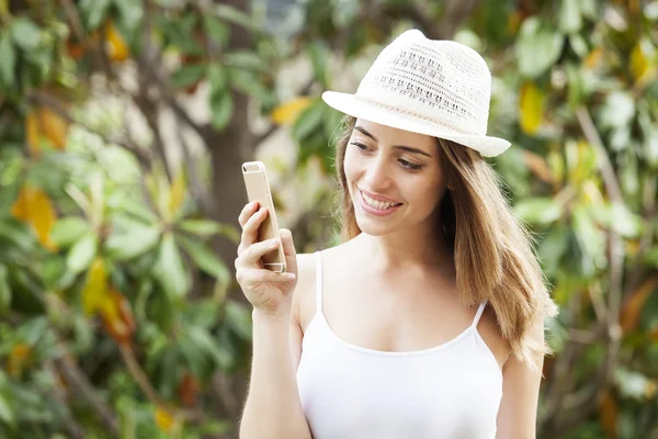 Portrait of a smiling young woman wearing a hat and standing in a garden looking at her cell phone.