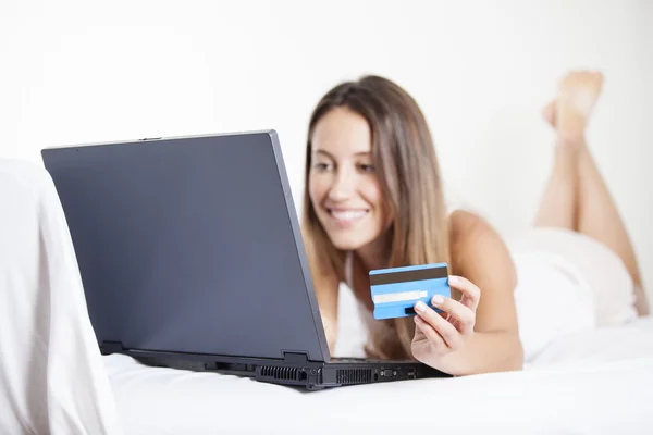 A smiling woman on her bed with laptop in front of her and credit card in hand