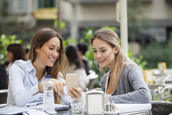 Two happy friends or sisters sharing a smart phone in a coffee shop terrace