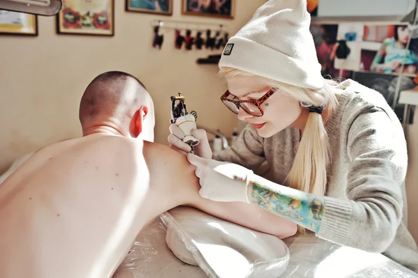 Young blonde woman master tattooist in glasses and hat with tatt