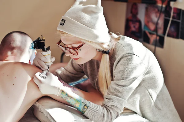Young blonde woman master tattooist in glasses and hat with tatt