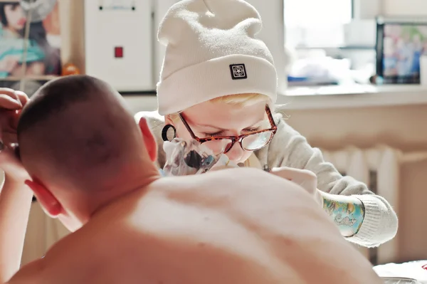 Young blonde woman master tattooist in glasses and hat with tatt