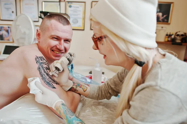 Young blonde woman master tattooist in glasses and hat with tatt