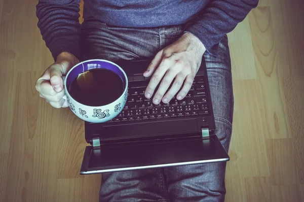 Young student with a cup of tea using a laptop and sitting on the wooden floor in a classroom