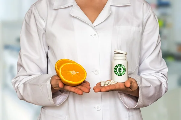 Young doctor holding two halves of orange and bottle of pills with vitamins and compare them