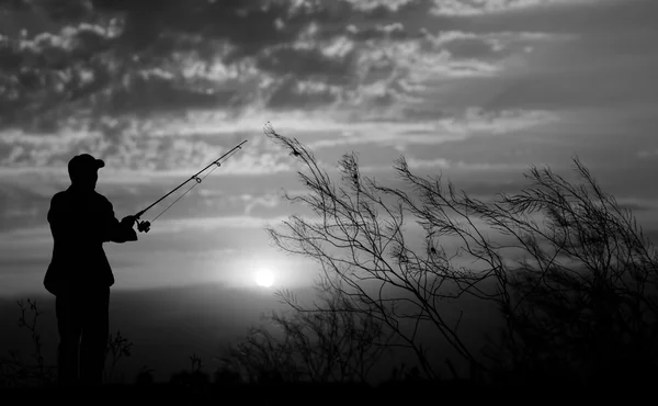 Fisherman standing with fishing rod near river on background of plants and beautiful cloudy sky with sunset and rays of light black and white