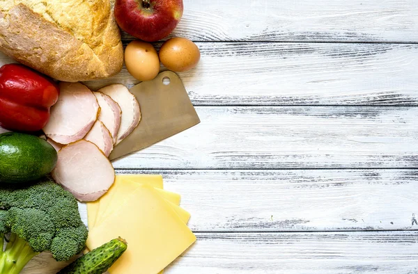 Set of products for cooking - apple, bell pepper, cucumber, fresh bread, eggs, sliced ham, broccoli, cheese, avocado and cleaver on white vintage wooden work surface