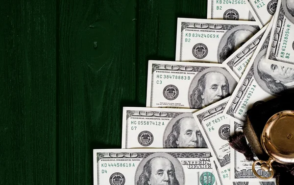Stack of money dollars laid out like a ladder with antique gold watch on dark green retro stylized wood background