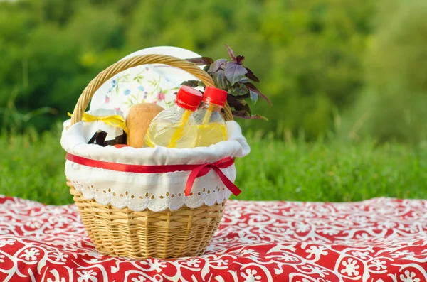 Decorated picnic basket, plate, bun, jar of jam, lemonade, bunch of basil, on red tablecloth, green landscape