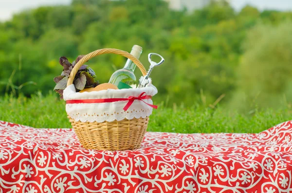 Decorated picnic basket with a bottle of white wine, corkscrew, buns and bunch of basil on red tablecloth, green landscape