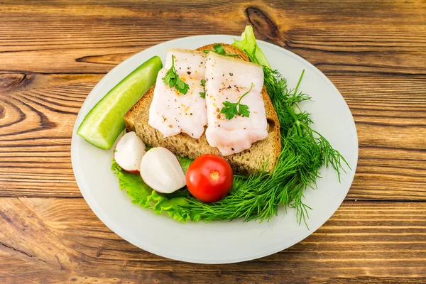 Sandwich with slice of rye bread, fresh pork lard and parsley, fresh produce, tomato in the plate on wooden table.