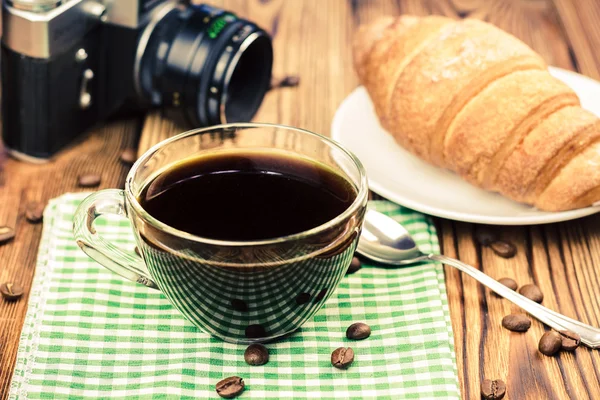 Cup of black coffee on green napkin with croissant, vintage camera, wooden table in cafe. travel concept