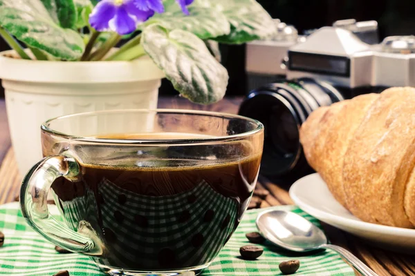 Cup of black coffee on green napkin with croissant, vintage camera, flower pot, wooden table in cafe. travel concept