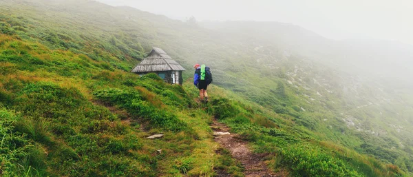 Tourist goes along mountain trail to the small wooden house on hill of ridge in mist, Carpathians, Ukraine.
