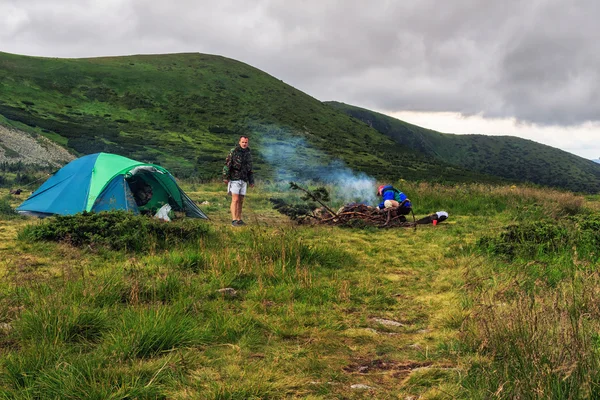 Camping tent and two men tourists at campfire in Carpathian mountains, summertime journey.