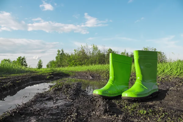 Green rubber boots in the mud next to a puddle on a wet country road