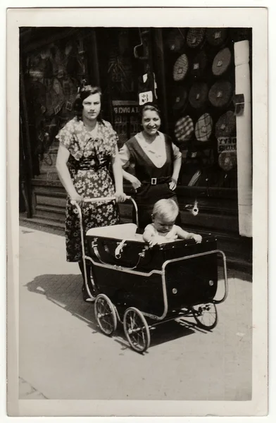 A vintage photo shows women go for a walk with pram (baby carriage). Antique black & white photo.