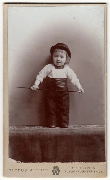 Vintage cabinet card shows cute small boy wears bowler hat, bow tie, cummerbund and holds walking stick. Antique black & white photo.
