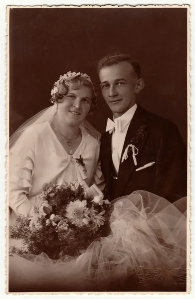 Vintage photo of newlyweds with wedding bouquet. Bride wears wedding veil headdress. Groom wears posh clothing, white bow-tie. Black & white antique studio portrait.
