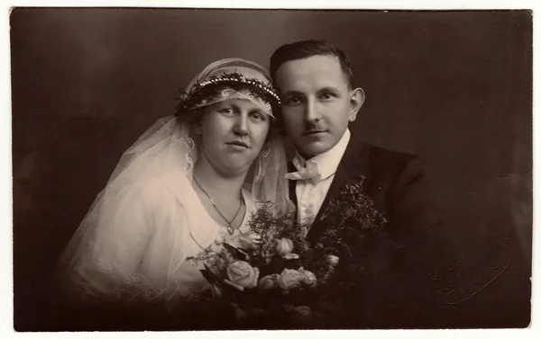 Vintage photo of newlyweds with wedding bouquet. Bride wears wedding veil headdress. Groom wears posh clothing, white bow-tie. Black & white antique studio portrait.