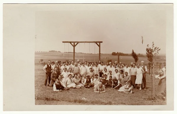 Vintage photo shows a group of people outdoors. Horizontal bar for rope- climbing is in the background.  Black & white photo.