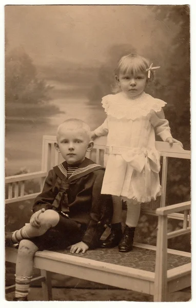 Vintage photo shows two children (siblings). Boy wears sailor costum and girl wears white dress, laced collar and hair ribbon. They pose on historic white bench. Black & white studio photo.
