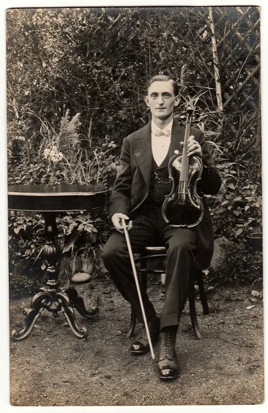 Vintage photo shows young man poses with the violin outdoors. The violinist sits at the black elegant table. Antique black & white photography.