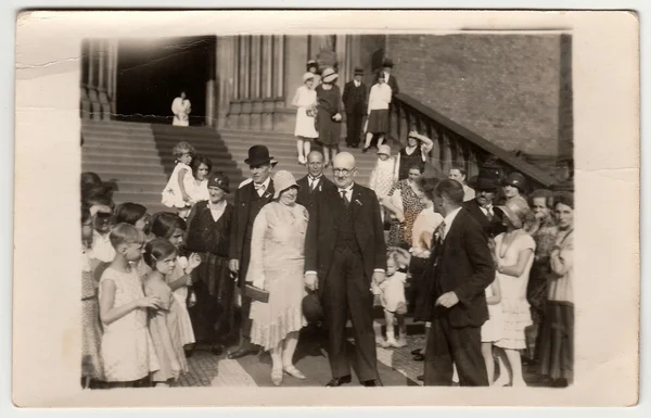 Vintage photo shows elderly newlyweds in front of church after wedding ceremony. Black & white antique photography.