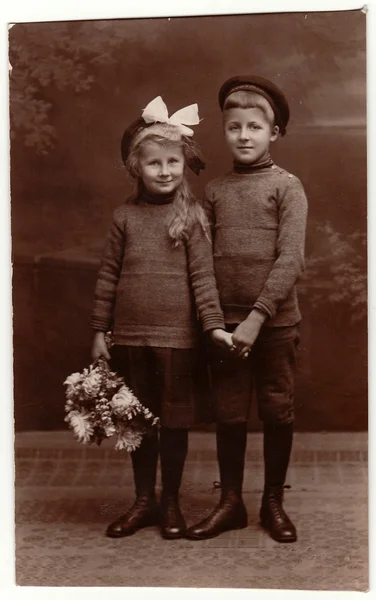 Vintage photo shows girl and boy (siblings), girl wears white hair ribbon. Black & white studio photography.